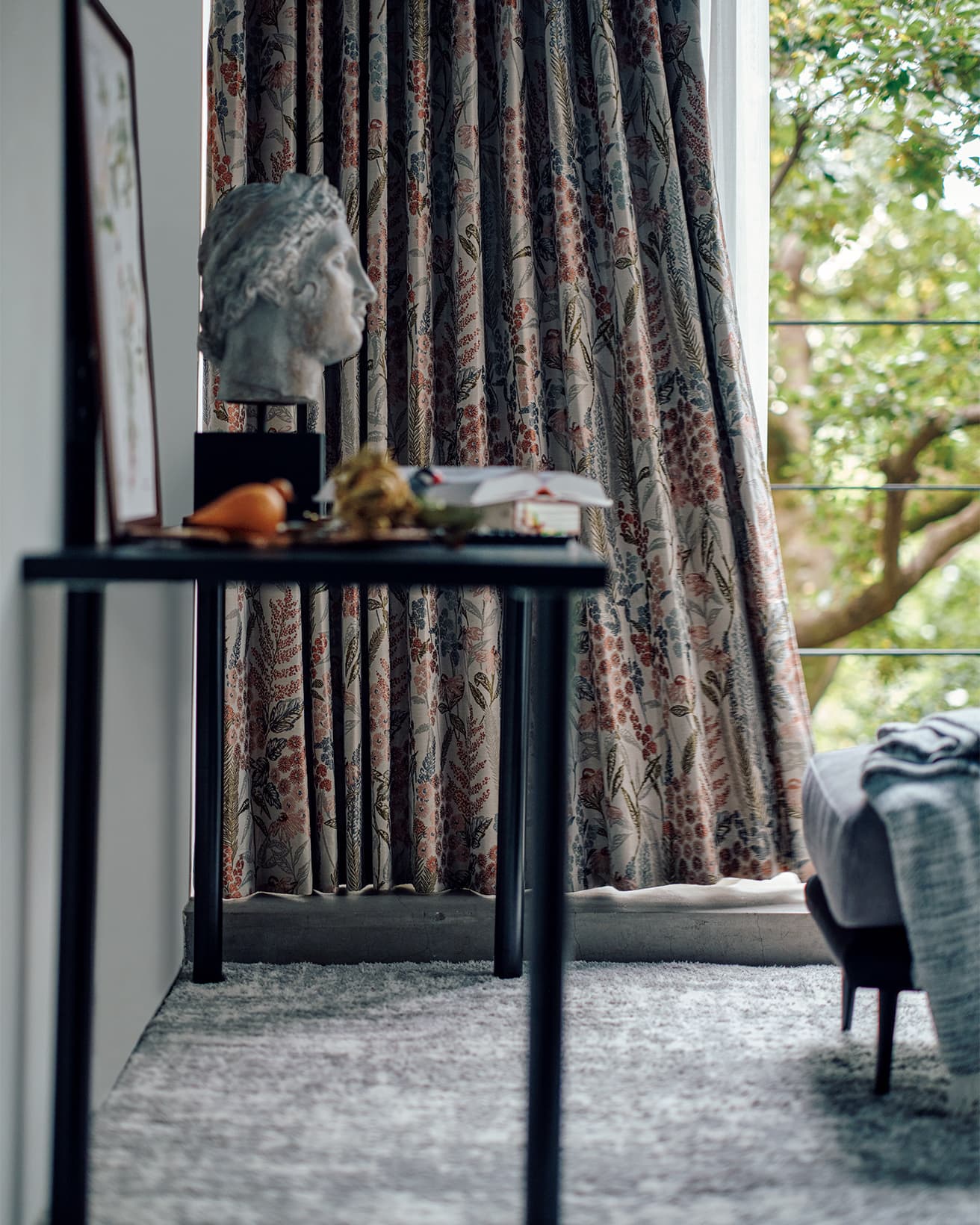 同じ柄のカーテンがさまざまな雰囲気の空間で美しい暮らしを演出してくれるのも、ジャカードカーテンならではの魅力といえる。オーダーカーテン1.5倍ヒダ（Ｗ176×Ｈ213㎝×２枚）30,900円、オーダーレースカーテン２倍ヒダ（Ｗ176×Ｈ212㎝×２枚）33,900円、フリーカバー2,990円、スツール（Ｗ60×Ｄ50×Ｈ43㎝）19,990円、アクセントラグ（約133×190㎝）17,990円、組み合わせデスク各種：天板（Ｗ140×Ｄ59×Ｈ３㎝）7,000円、脚2Pセット（２個使用）（Ｗ９×Ｄ９×Ｈ69㎝）2,000円（以上ニトリ）。テーブルなどに置かれた彫像、書籍、置物などはスタイリスト私物。